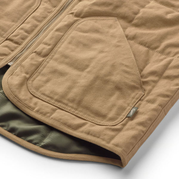 Close-up of a tan quilted jacket with a large front patch pocket, zipper closure, and a small metal brand tag, shown on a white background.