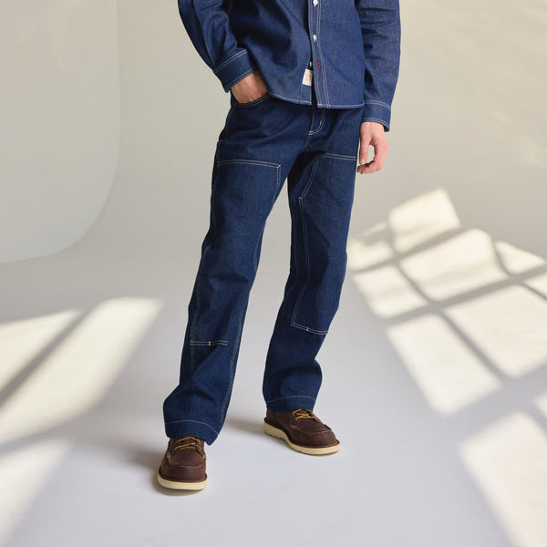 Person wearing blue jeans and a denim shirt standing in a room with sunlight casting shadows on the wall.
