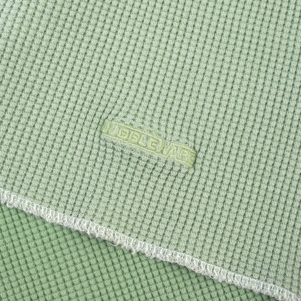 Close-up of a light sage green waffle-weave fabric with a white frayed hem and a small tonal embroidered 'WUBLEWARE' brand tag.