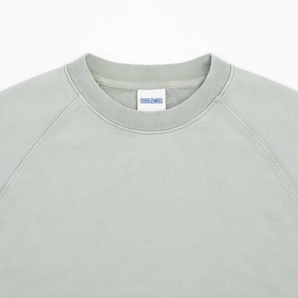 Close-up of a crewneck sweatshirt in a light sage green color, showing the ribbed collar and a white neck label with blue text.