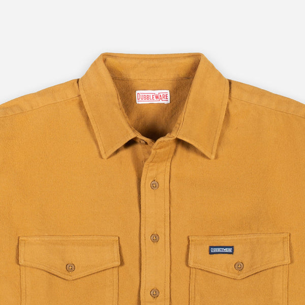 Close-up of a mustard yellow button-up shirt with a pointed collar, two chest pockets, and a visible Dubbleware neck label and a small chest patch logo.
