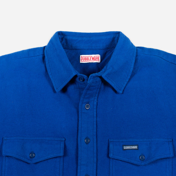 A close-up of a royal blue button-down shirt by Doubleware, featuring a classic collar, dark buttons, and two buttoned chest pockets on a light gray background.