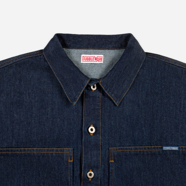 Close-up of a dark indigo denim button-up shirt by Dubble Wade, showing the collar, front buttons, chest pockets, and a small brand tag on the right chest pocket.