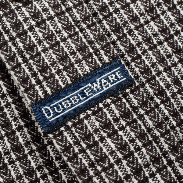 Close-up of a woven knit fabric with a dark brown and off-white textured pattern, featuring a blue embroidered label with the brand name 'OUBLEWARE' in white.
