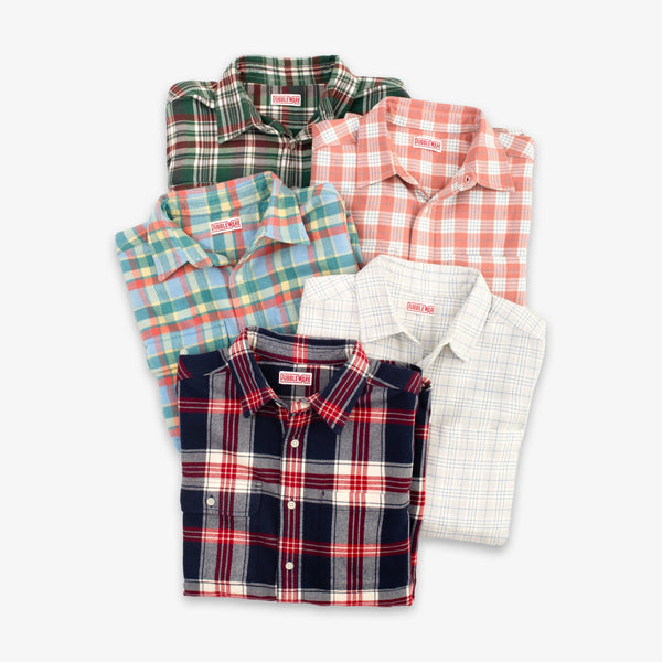 Five folded plaid button-up shirts in different colorways arranged in a fan shape on a light gray background. The shirts include green, coral, blue, red, and white plaid patterns.
