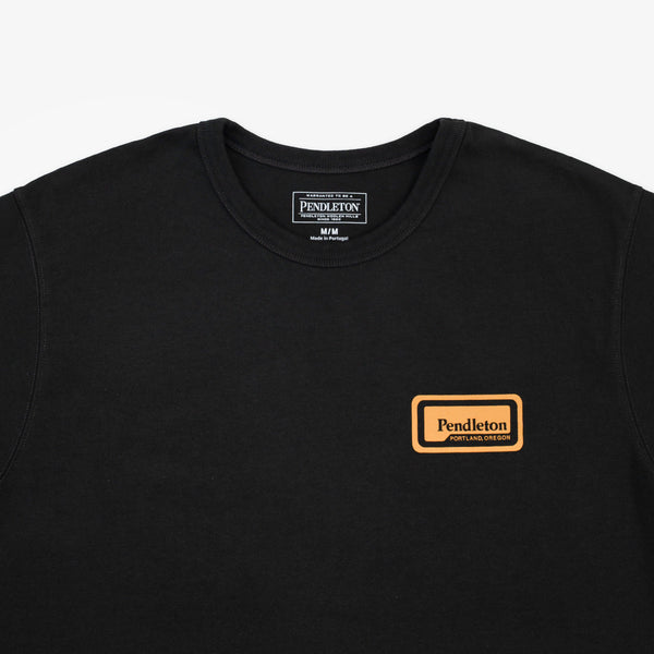A close-up of a black Pendleton crewneck sweatshirt showing the chest area, featuring a small tan and black embroidered logo patch on the left chest and a larger brand label inside the collar.