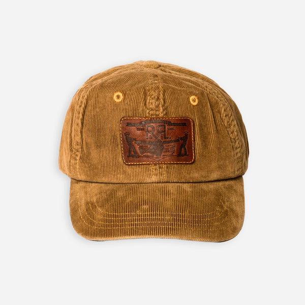 A brown corduroy baseball cap with a brown leather patch on the front featuring a stylized eagle logo, photographed against a light gray background.