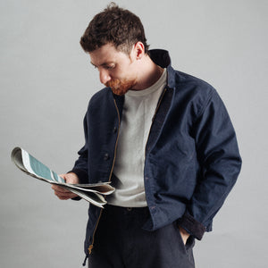 Double RL By Ralph Lauren - RRL Bower Deck Jacket Navy - A man with a beard looking down at a magazine, wearing a navy blue overshirt over a light beige t-shirt and dark trousers, against a plain gray studio background. - Alternative View 1