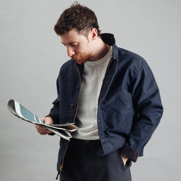 A man with a beard looking down at a magazine, wearing a navy blue overshirt over a light beige t-shirt and dark trousers, against a plain gray studio background.