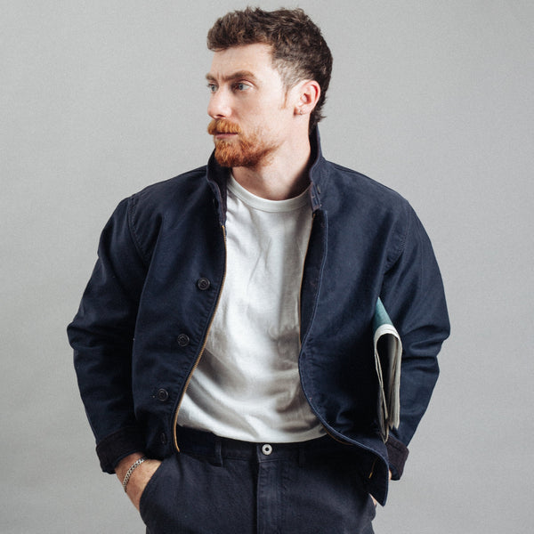 A man with a reddish beard wearing a dark navy blue corduroy jacket over a white t-shirt and dark trousers, holding a notebook in his pocket against a plain gray studio background.