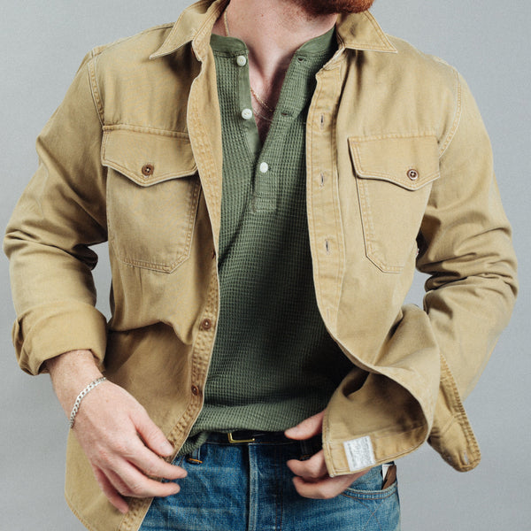 A man wearing a tan utility overshirt over a green henley shirt and blue jeans, shown from the neck down against a plain gray studio background.
