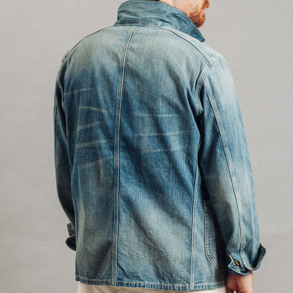 A man wearing a light blue washed denim jacket, shown from the back, paired with off-white trousers against a neutral gray studio background.