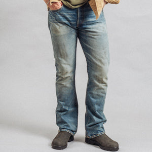 Double RL By Ralph Lauren - RRL Slim Boot Selvedge Jeans Briarfield Wash - A person wearing light-wash blue denim jeans with a tan overshirt and dark brown boots, shown from the waist down against a plain light gray studio background. - Alternative View 1