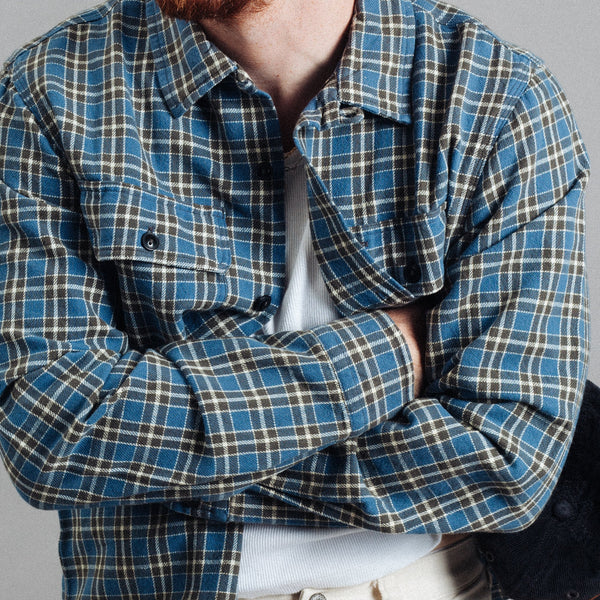 A person wearing a blue, green, and beige plaid button-up shirt over a white undershirt, paired with off-white pants, shown from the neck down against a plain gray background.