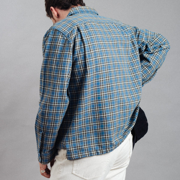 A man wearing a blue and beige plaid flannel shirt, shown from the back, paired with white pants against a plain gray background.