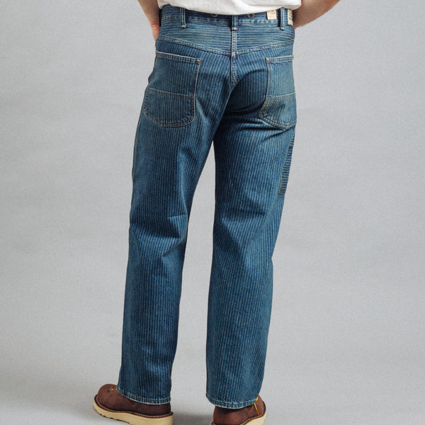 A person shown from the back wearing blue denim jeans, a white t-shirt, and brown shoes against a plain gray studio background.
