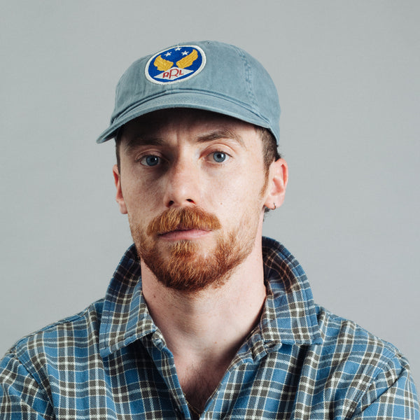 A man with a red beard and blue eyes wearing a light blue baseball cap with a circular 'HPL' logo patch and a blue and green plaid shirt, photographed against a solid gray background.