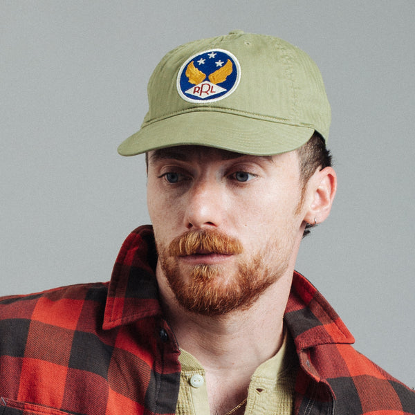 A man wearing an olive green baseball cap with a circular 'APL' eagle patch on the front, paired with a red and black plaid flannel shirt, photographed against a solid gray background.