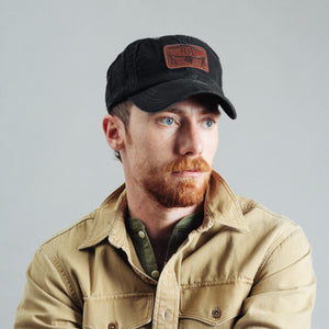 Double RL By Ralph Lauren - RRL Leather Patch Corduroy Trucker Cap Black - A man with a red beard wearing a black baseball cap with a brown patch and a tan button-up shirt, photographed against a plain light gray background. - Alternative View 1
